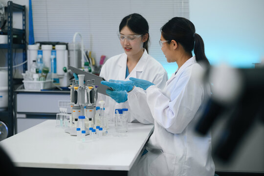Two Asian Female Scientists Reviewing Data on Tablet in Modern Laboratory with Pipettes and Sample Vials