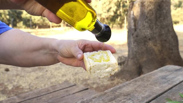 Close up of a person's hands pouring golden extra virgin olive oil from a bottle onto a slice of bread. Healthy mediterranean diet food tasting