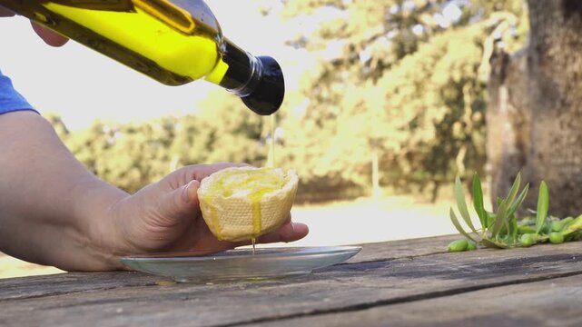 Female hands pouring golden extra virgin olive oil from a bottle onto a piece of fresh bread over a wooden table in a rural setting