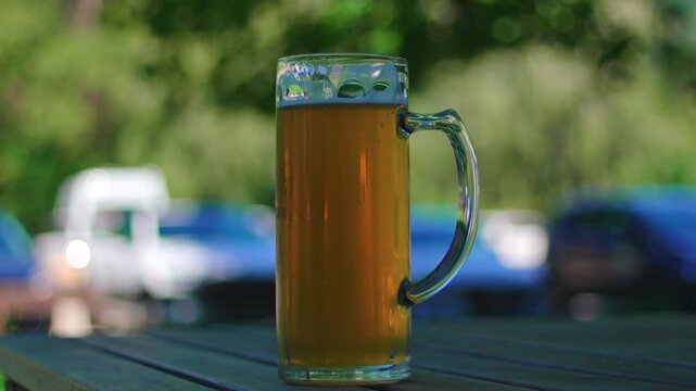 Close view of cold beer mug on outdoor picnic table with blurred cars and green landscape in background conveying summer relaxation casual dining and refreshment