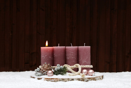 First Sunday of Advent: A rustic Advent wreath in the snow with a pink-glowing pillar candle on a tree stump. In the background, a wooden wall with a copy room.
