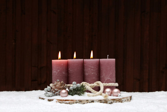 Third Sunday of Advent: A rustic Advent wreath in the snow with three pink-flame pillar candles on a tree stump. In the background, a wooden wall with a copy room.