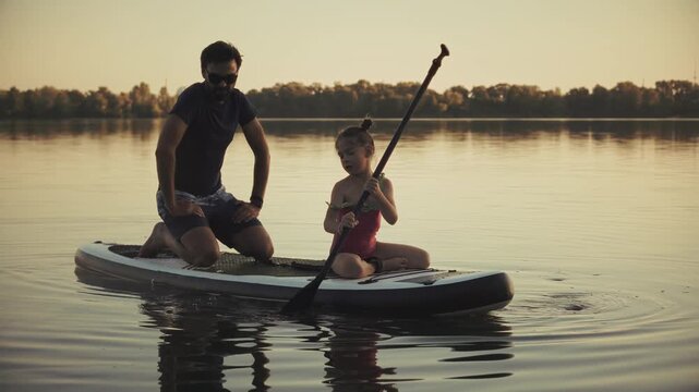 Father Watching Daughter Holding Oar And Sitting On Inflatable Stand Up Paddleboard Floating On Calm Water At Sunset