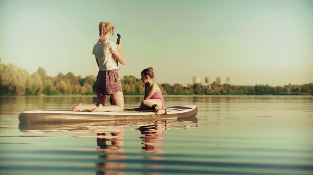 Mother And Child Sitting On Inflatable Sup Board Floating On Calm Water River 