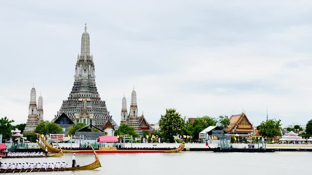 A golden Thai royal barge processional rowing past the iconic Wat Arun temple on the Chao Phraya River in Bangkok, Thailand during a traditional cultural ceremony under a bright daylight sky.