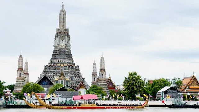 A golden Thai royal barge processional rowing past the iconic Wat Arun temple on the Chao Phraya River in Bangkok, Thailand during a traditional cultural ceremony under a bright daylight sky.