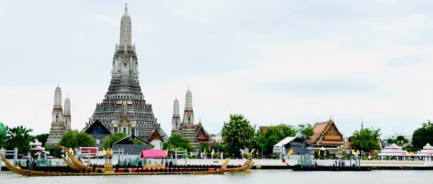 A golden Thai royal barge processional rowing past the iconic Wat Arun temple on the Chao Phraya River in Bangkok, Thailand during a traditional cultural ceremony under a bright daylight sky.
