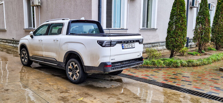 A white Radar RD6 electric pickup truck with a Georgian license plate is parked in front of a building; a modern Chinese electric pickup truck from Geely is driving on the road near Batumi, Georgia
