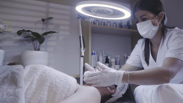 Masked esthetician gently massaging client face under ring light