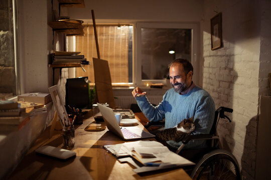 Smiling man in wheelchair working on laptop at home office with cat
