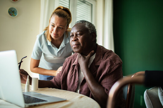 Senior man with caregiver consulting online doctor at home on laptop