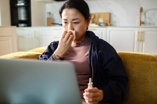 Sick woman using tissue and nasal spray while using a laptop at home