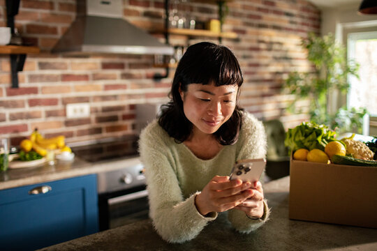 Woman using smartphone in home kitchen with fresh groceries