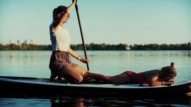 Mother Kneeling With Oar While Daughter Lies Down On Stand Up Paddleboard Floating On Calm River Water 