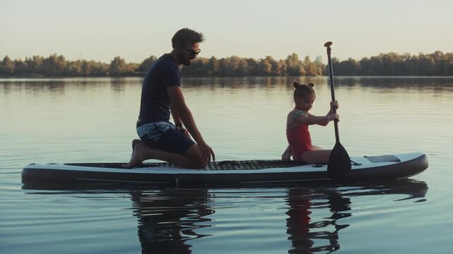Man Kneeling On Stand Up Paddle Board Watching Child Paddling On Calm Lake Water 