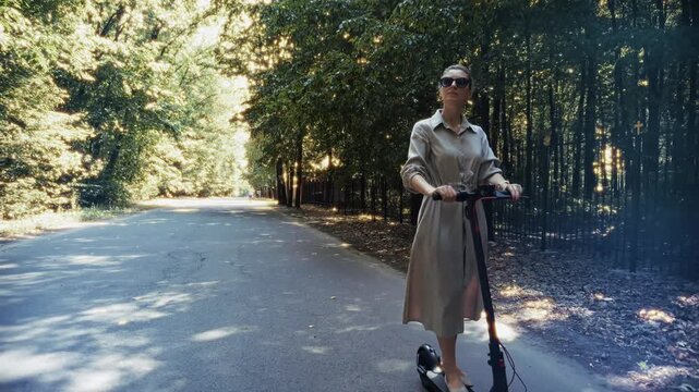 Woman Riding Electric Kick Scooter Along Asphalt Road In Green Forest Park 
