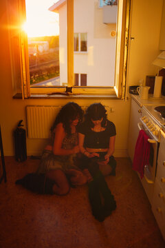 Fashionable lesbian couple using digital tablet sitting by window in kitchen at home