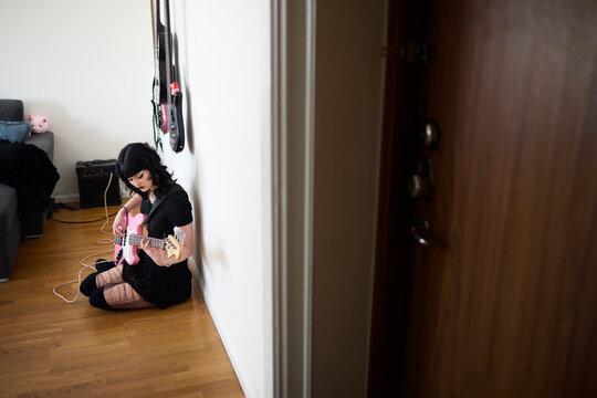 Young woman playing electric guitar connected to amplifier while sitting on floor at home