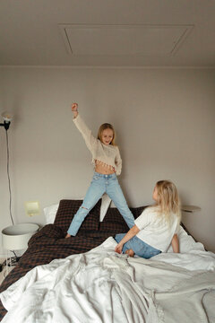 Girl sitting on bed while looking at sister dancing in home
