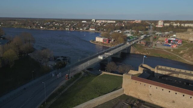 An aerial view of the Friendship Bridge and border checkpoint over the Narva River connecting Narva, Estonia, and Ivangorod, Russia, with the Estonian flag flying on Narva Castle in the foreground.