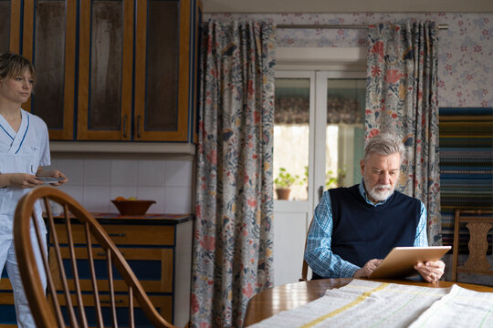 Female nurse bringing breakfast for senior man using digital tablet while sitting at table in home