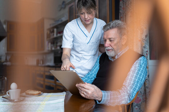 Female caregiver teaching elderly man to use digital tablet while sitting for breakfast at home