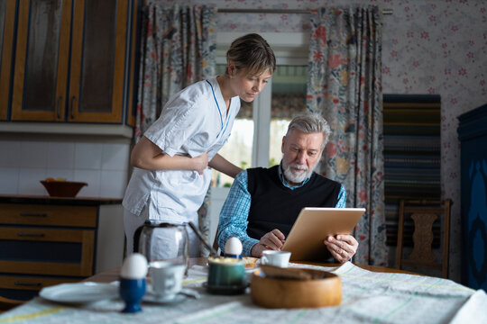 Female caregiver looking at digital tablet used by elderly man sitting for breakfast at dining table in home