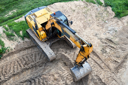 Yellow crawler excavator stands on sand at construction site, top-down view, aerial photography, industrial building machinery works with ground, bucket digs soil on earthworks area