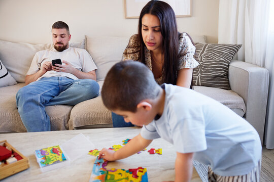 Man using smart phone while woman assisting son making jigsaw puzzle at home