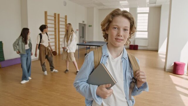 Portrait of Caucasian teenage boy holding book and wearing backpack in school gym while camera zooming in. Footage is perfect for high school education and student life marketing campaigns