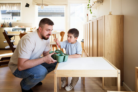 Man crouching near boy choosing colors from container while sitting at table in home