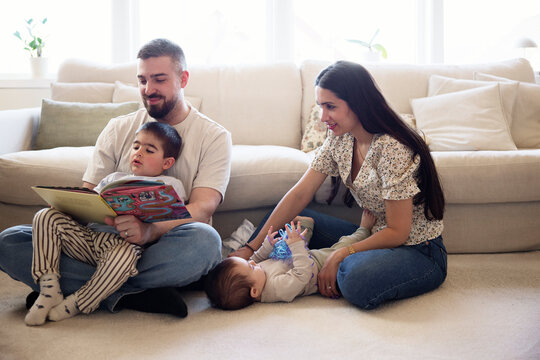 Woman sitting with toddler beside man and son reading picture book near sofa in living room at home