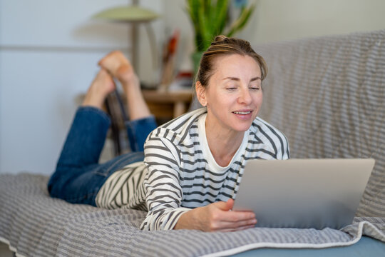 Relaxed carefree woman resting on couch at home with laptop, watching media and shopping online, smiling cheerful female comfortably lying on sofa browsing online on computer, lazy leisure time.