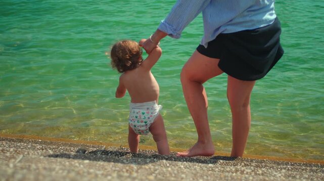 Mother guiding toddler down lakeside steps into shallow water during sunny outdoor outing capturing safety, confidence building and authentic summer recreation