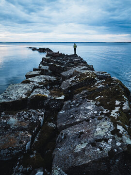 Wide path of boulder rocks amidst sea water with unrecognizable person looking at view in distant under cloudy sky