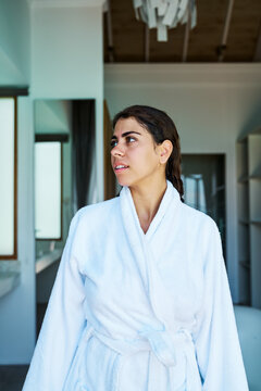 A woman wearing a white bathrobe looks thoughtfully indoors in a home