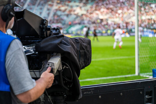 Television camera operator filming a professional soccer match from the sidelines, with a broadcast camera in the foreground and football players on the field.