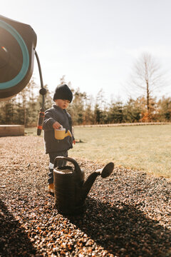 Cute boy standing with small watering can on gravel surface in back yard at sunny day