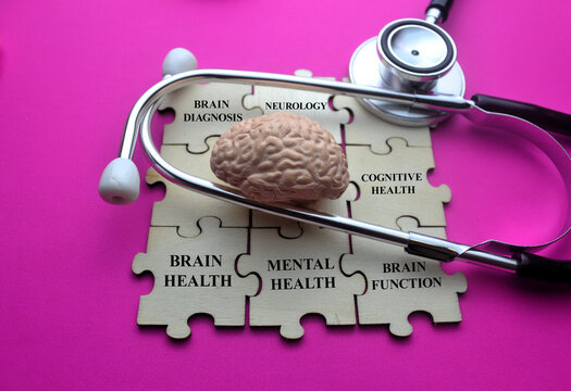 Stethoscope placed around brain model on wooden puzzle pieces labeled brain diagnosis neurology cognitive health mental health and brain function on pink background symbolizing neuroscience concept