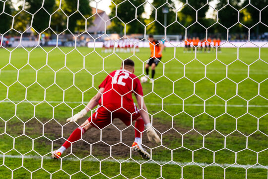 View through the net of a soccer goalkeeper in a red kit preparing to defend a penalty kick during a football match.