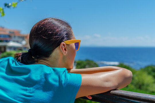 A woman in a blue shirt and yellow sunglasses leans on a balcony railing while looking out at the calm ocean view.