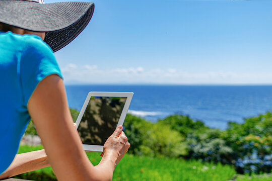 A woman wearing a black sun hat holds a digital tablet while sitting outdoors overlooking a beautiful blue sea.
