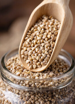 Buckwheat groats poured from a wooden scoop into a glass jar, emphasizing natural texture and whole grain detail. Neutral warm tones suit food packaging or healthy eating themes.