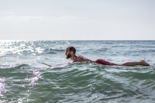 Male surfer paddles through rolling ocean waves while lying on his surfboard under bright summer light. Side-view action scene highlights endurance, training, and determination.