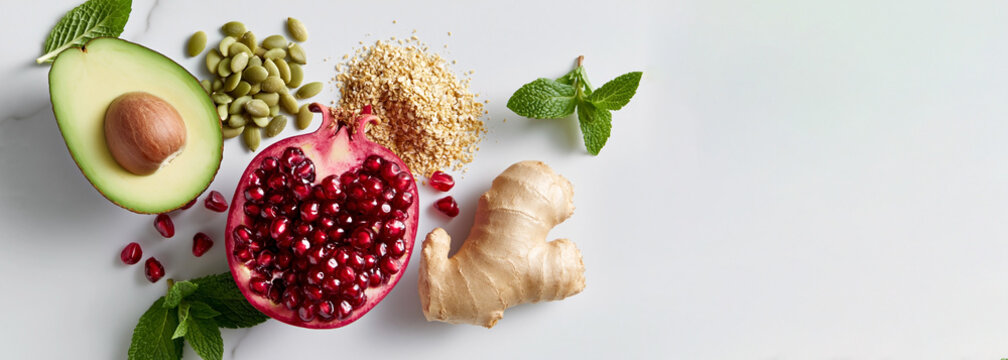 Fresh ingredients including avocado, pomegranate, ginger, mint leaves, and pumpkin seeds arranged on a light marble surface for healthy cooking or nutrition concepts