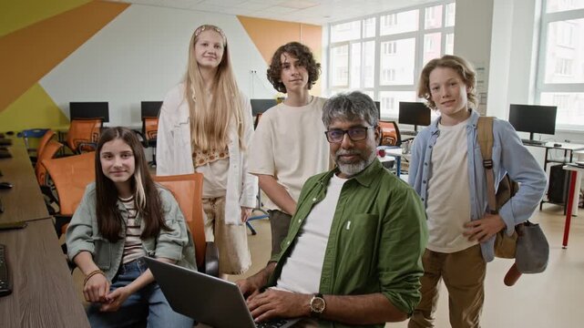 Portrait of middle aged Indian male teacher holding laptop and diverse teenagers looking at camera in computer class, zooming in. Video is perfect for promoting STEM education and school programs