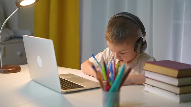 Kid wearing headphones writes in notebook doing schoolwork. Student focuses on homework tasks. Homework routine includes laptop work. Kid learns online with help of homework and student discipline.
