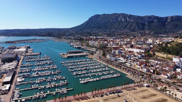 Aerial view of Denia marina with rows of moored yachts and boats, turquoise Mediterranean water, coastal town buildings, palm trees and Montgo mountain backdrop