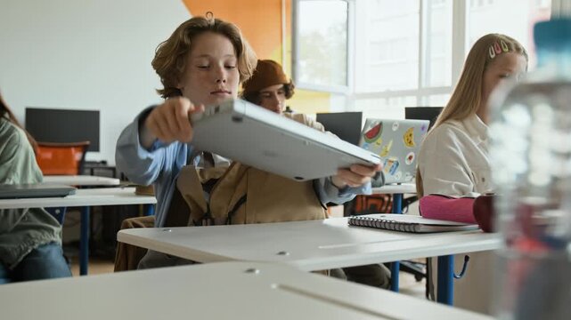Caucasian teenage boy taking laptop out of backpack and opening it on desk in classroom. Perfect for educational marketing, school technology promotions, and e-learning advertisements