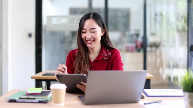 A smiling young Asian businesswoman wearing a red shirt sitting at a desk, looking at and working with a digital tablet and laptop in a modern office.